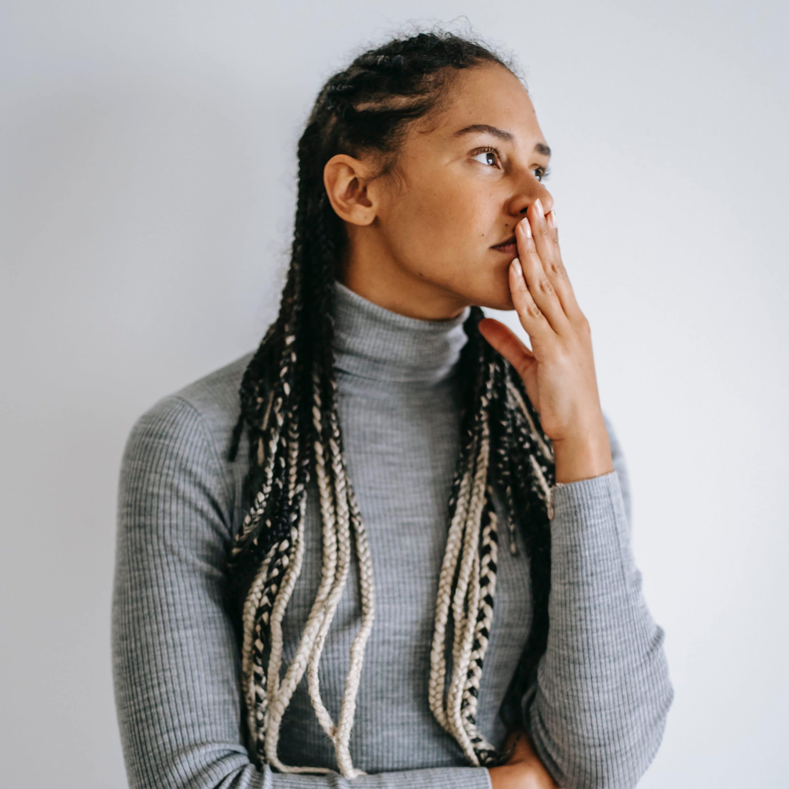 Anxiety can be soothed with sensory based practices. A woman with brown skin and long white-tipped braids anxiously touches her mouth while wearing a gray turtleneck sweater.