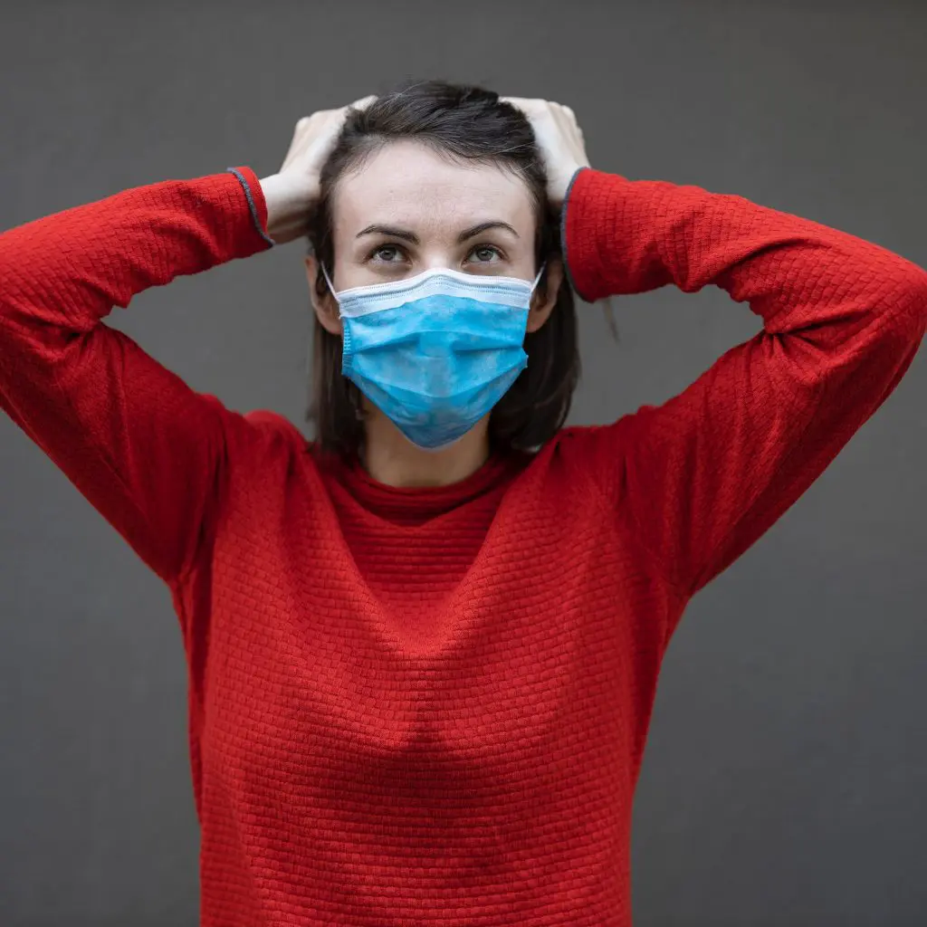 Stress surrounding COVID-19 can affect anyone. A woman wearing a red sweater and a surgical mask runs her hands through her hair while looking stressed.