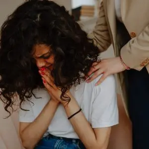 Grief can be a challenge for people of all ages. A young woman with curly dark hair cries into clasped hands while a female therapist pats her shoulders.