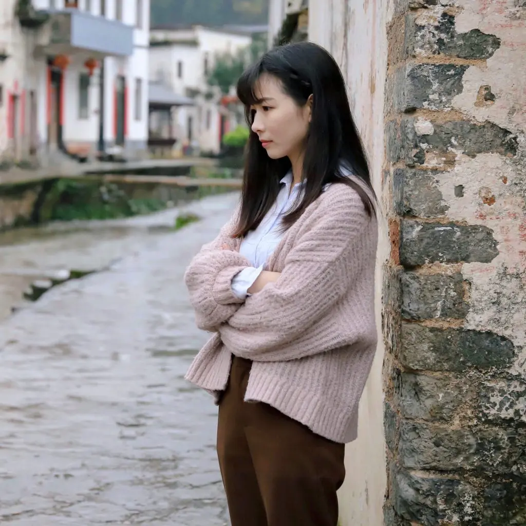 Trauma can stem from a number of experiences. A woman in a pink cardigan leans against a building with crumbling paint. 