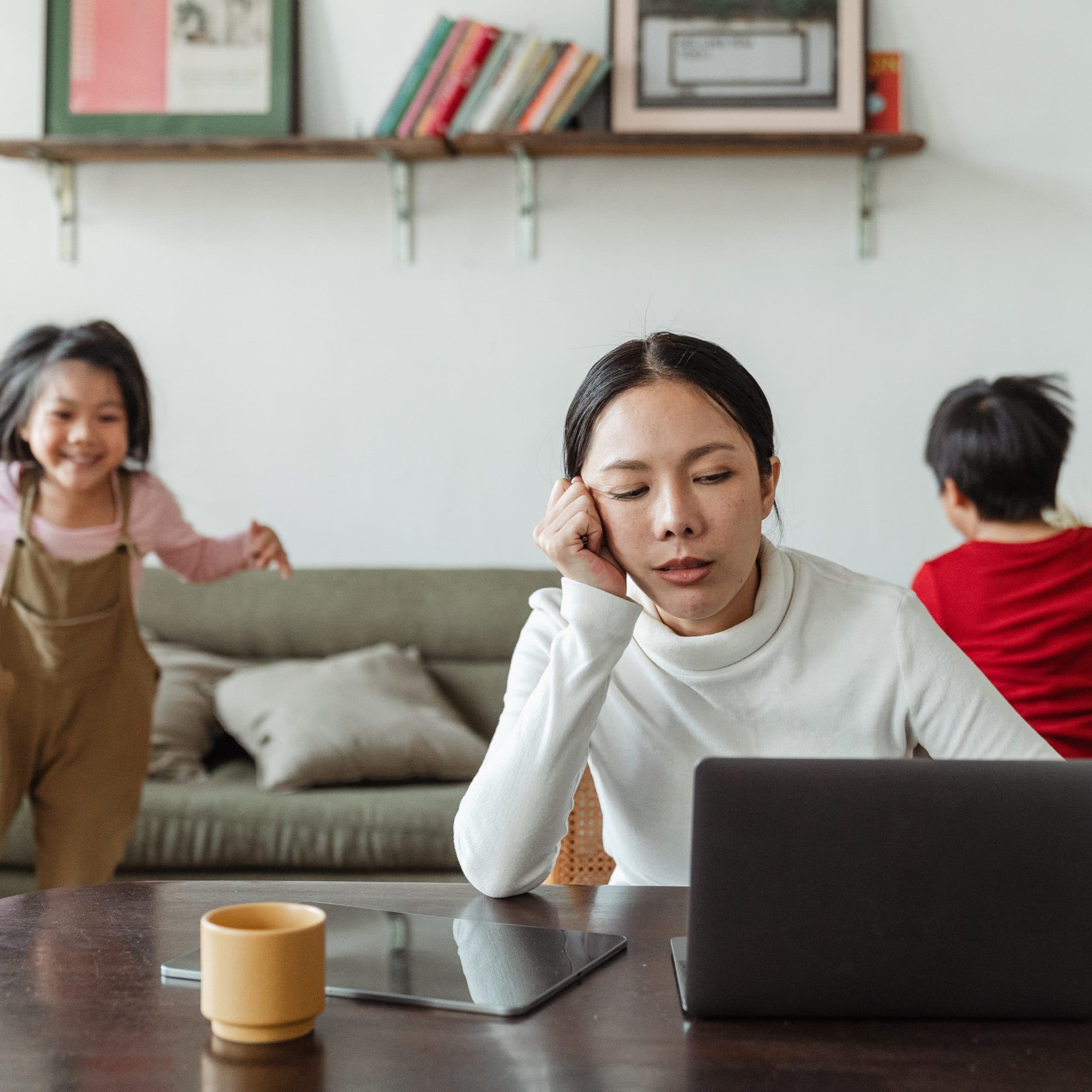 Stress can lead to feeling overwhelmed and burned out. A woman in a white sweater sits in front of her computer looking stressed while two small children play behind her.
