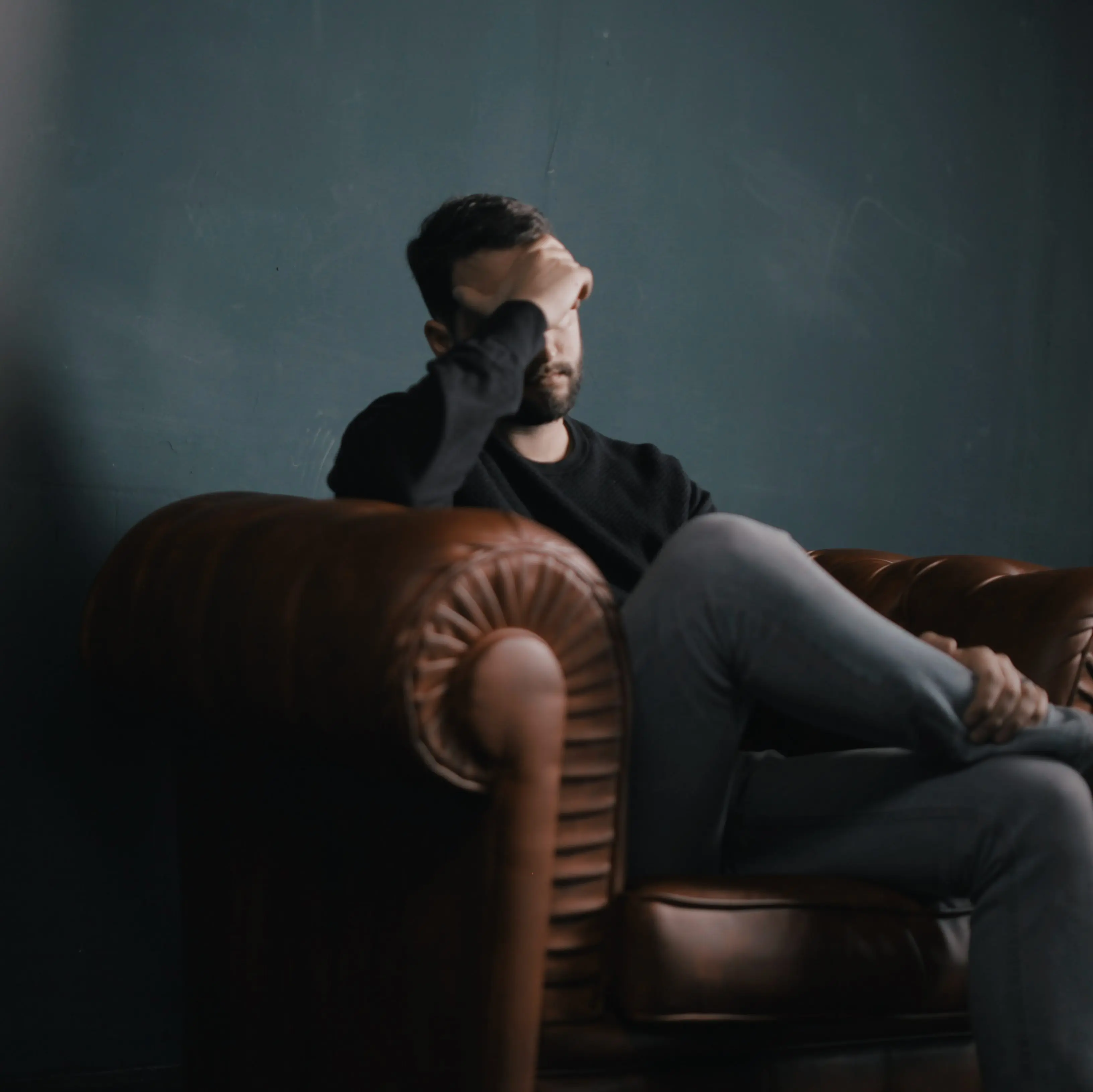 A man sits in a brown leather armchair and massages his temples.