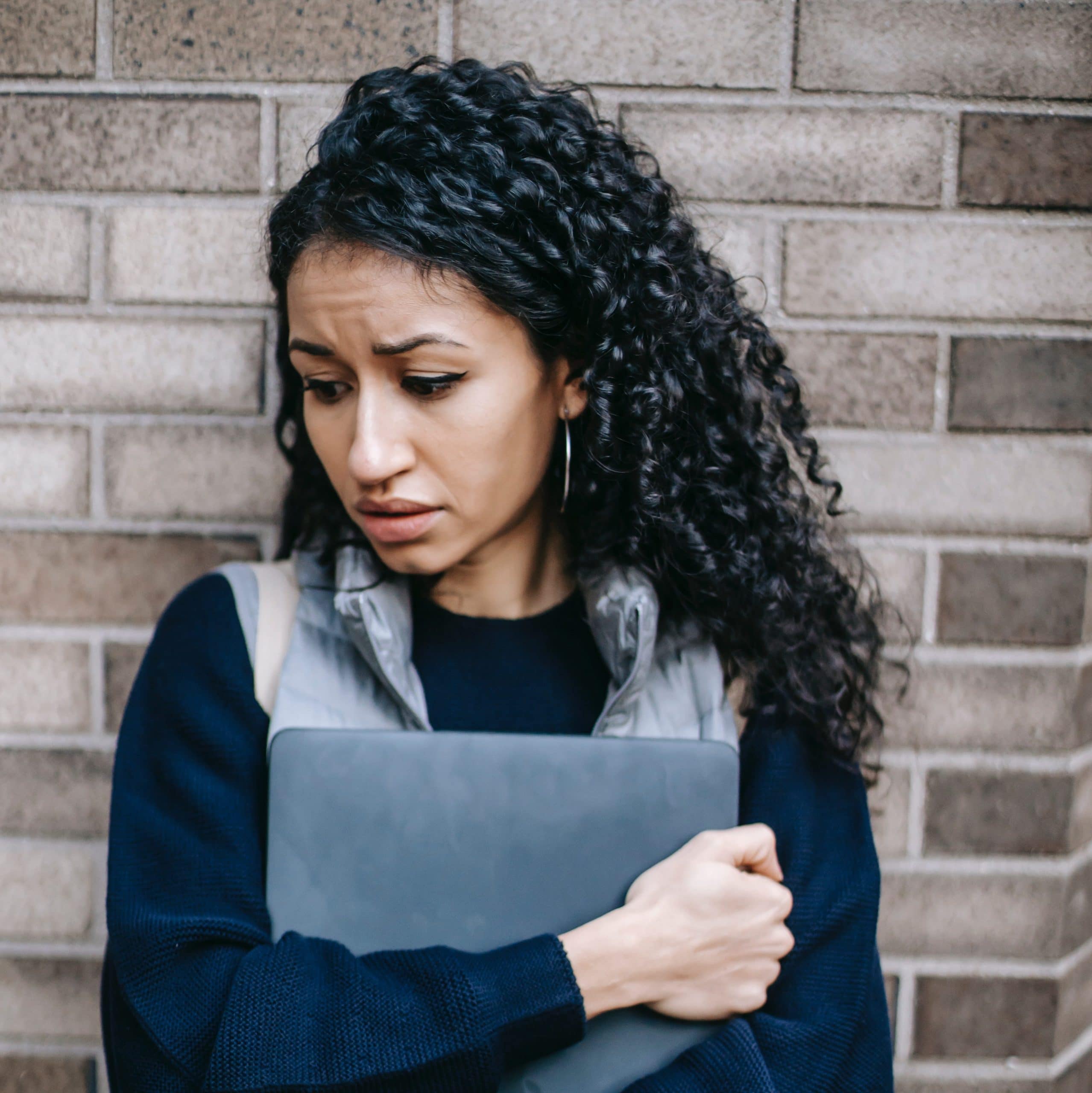 Trauma can affect anyone at any time. A Latina woman leans against a brick wall while holding a laptop and looking anxious.