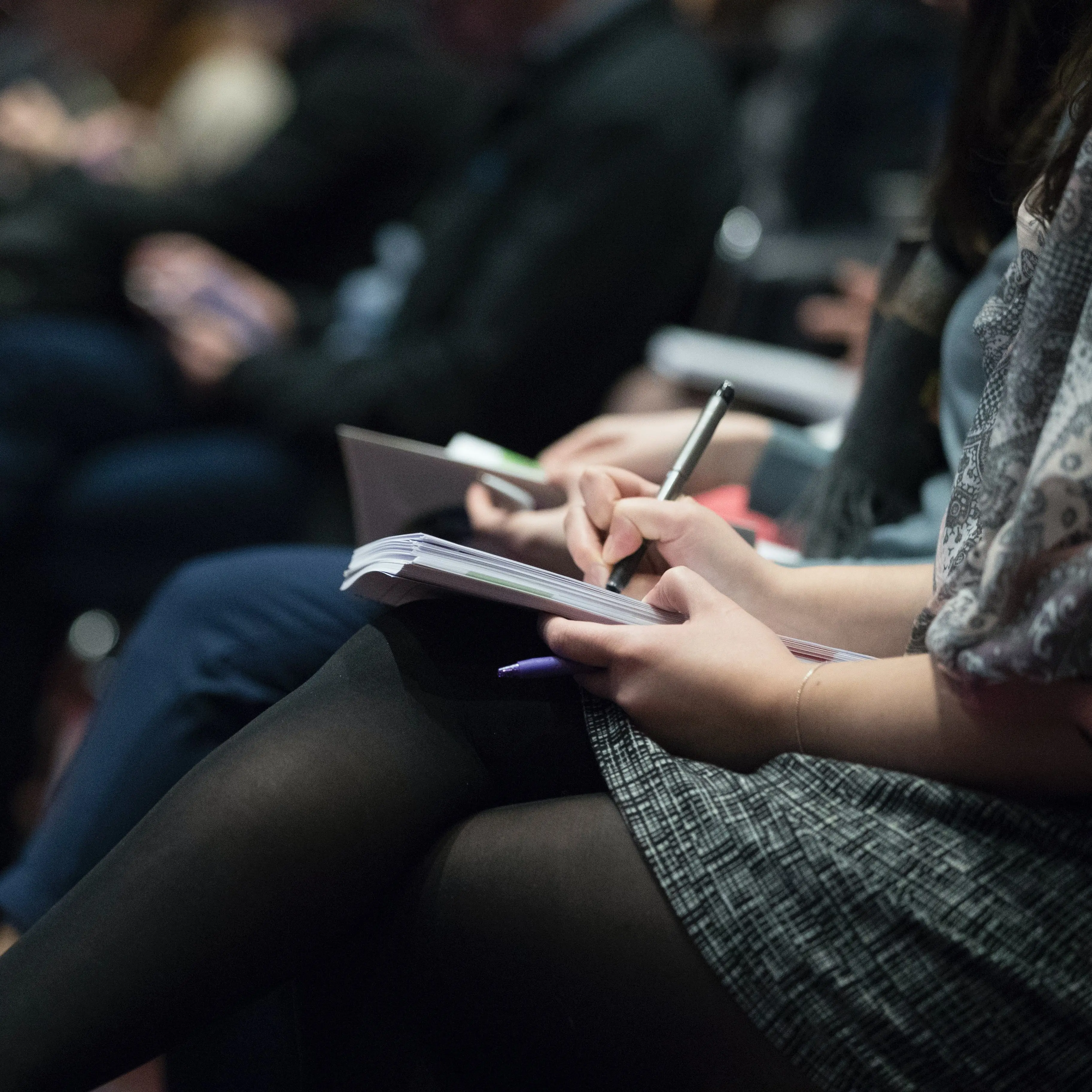 A person in the foreground wearing a skirt and tights takes notes while others in the background listen during a lecture.