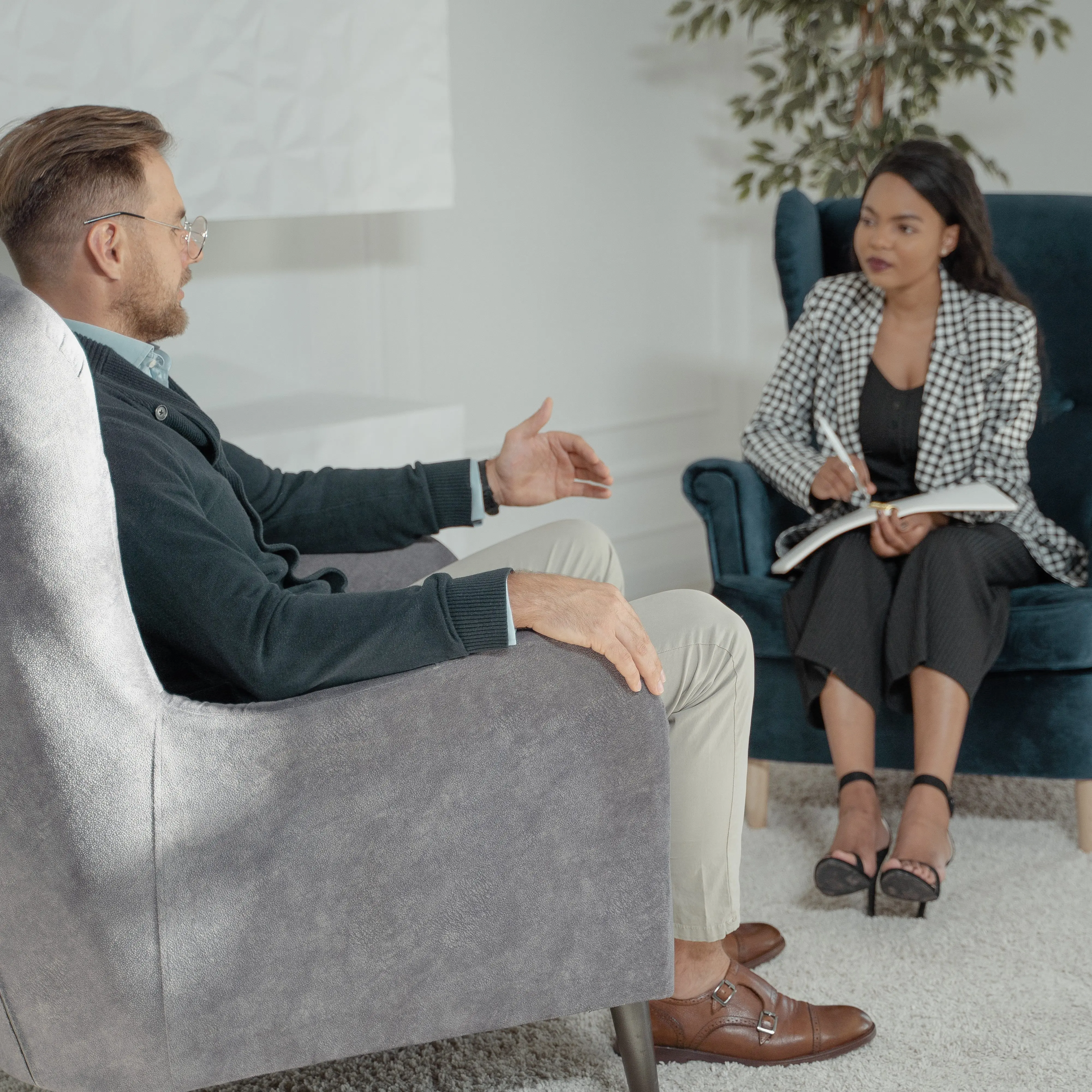A man and a woman in business clothing sit in wingback chairs and talk.
