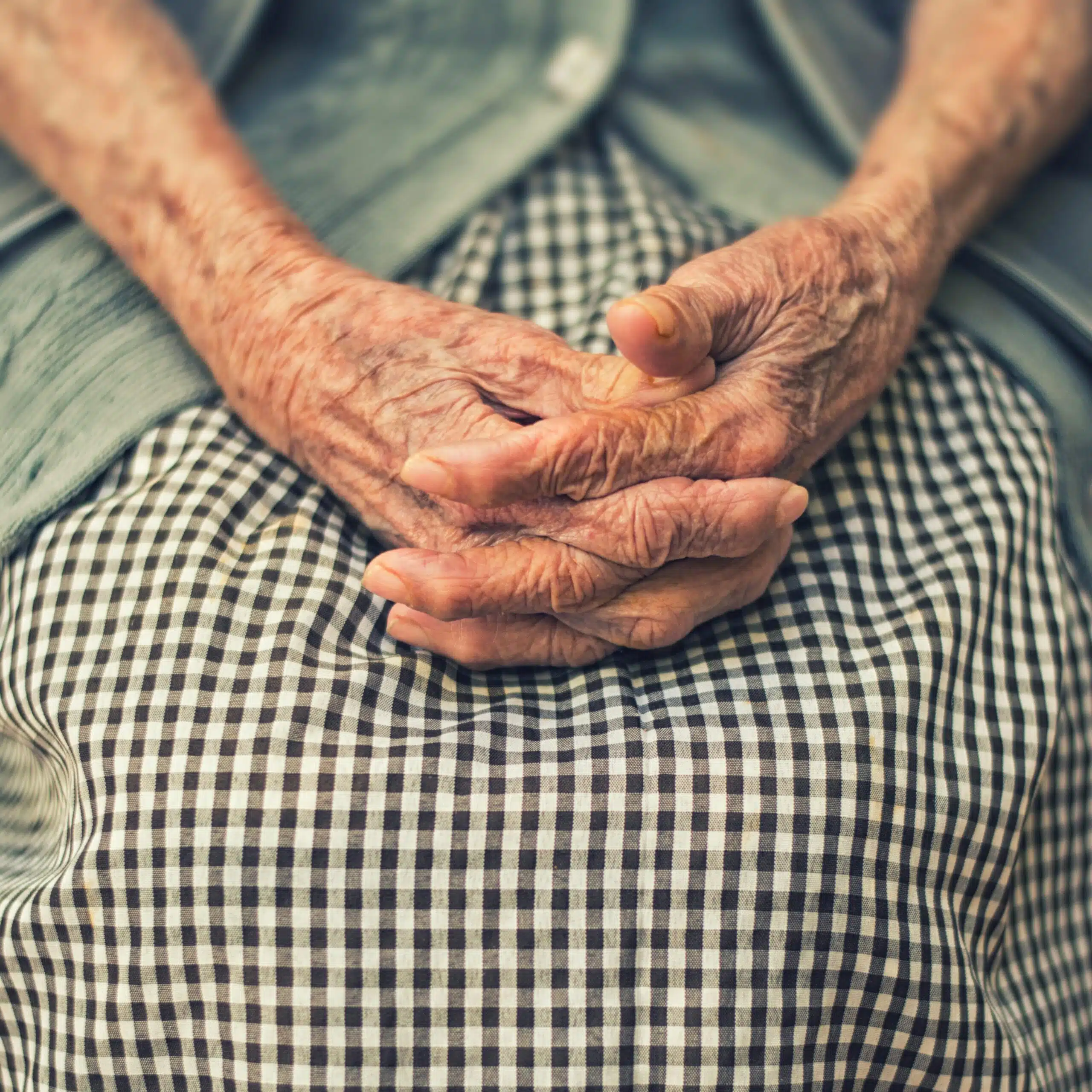 An older adult sits with their hands clasped in their lap while wearing a cardigan and plaid dress.